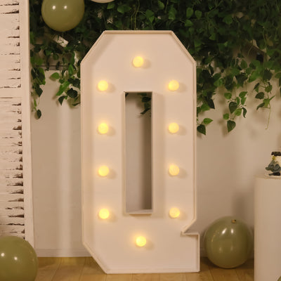 Illuminated marquee letter Q with warm lights, surrounded by greenery and balloons.