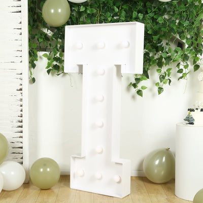 Large white marquee letter "T" with lights, surrounded by green foliage and balloons.