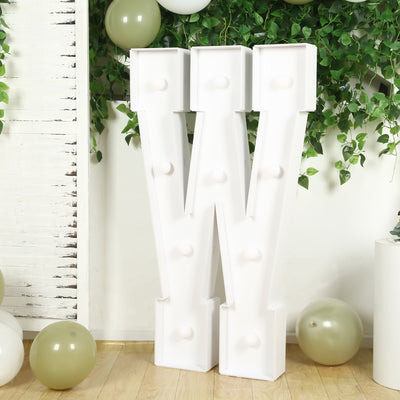 White marquee letter "W" with bulbs, set against greenery and balloons.
