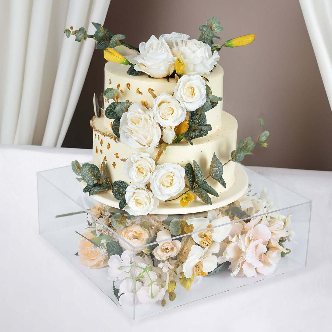 Wedding cake adorned with white roses and greenery in a clear display box.