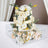Wedding cake adorned with white roses and greenery in a clear display box.