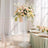 Elegant floral arrangement on a clear pedestal in a bright, airy setting.