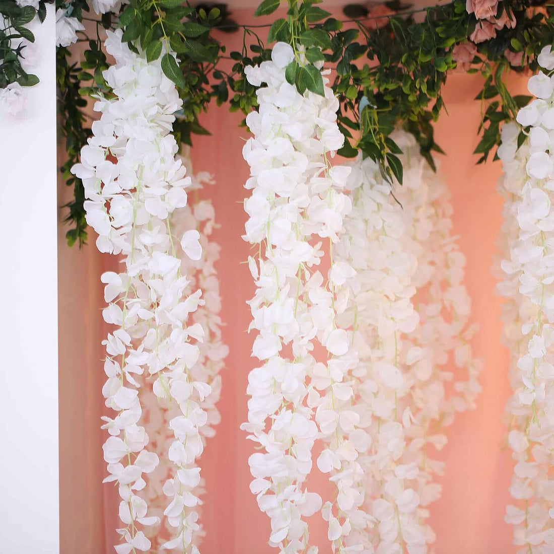 White floral hanging decor with greenery on a soft pink backdrop.