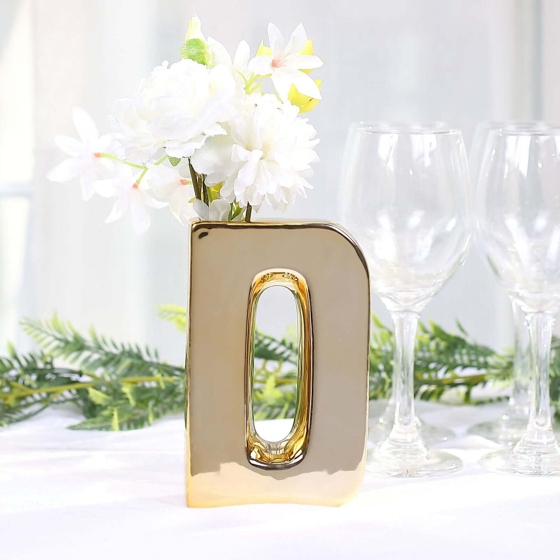 Gold letter "D" vase with white flowers and glasses in a bright setting.