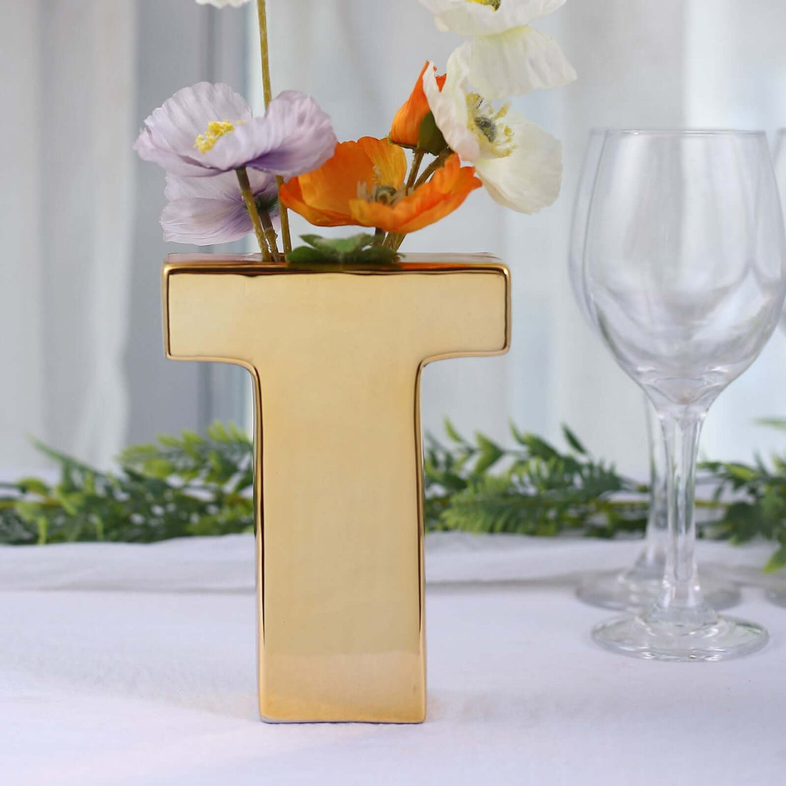 Gold letter "T" vase with colorful flowers and glassware in a stylish setup.