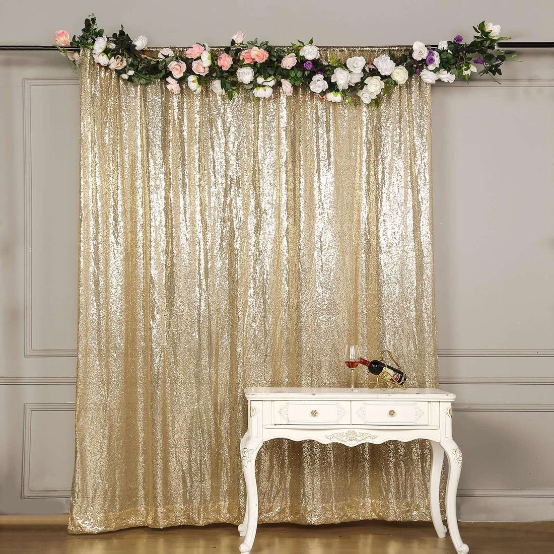 Gold sequin backdrop with floral garland and a white table with wine.