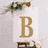 Gold glitter letter "B" on a decorated canvas with flowers.