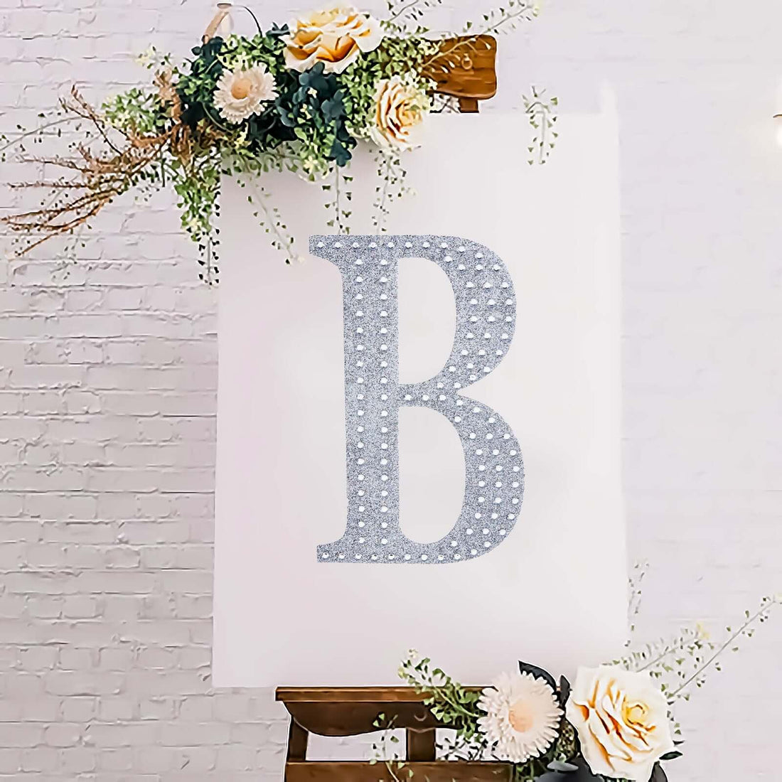 Glittery letter B on an easel surrounded by flowers in a stylish setting.