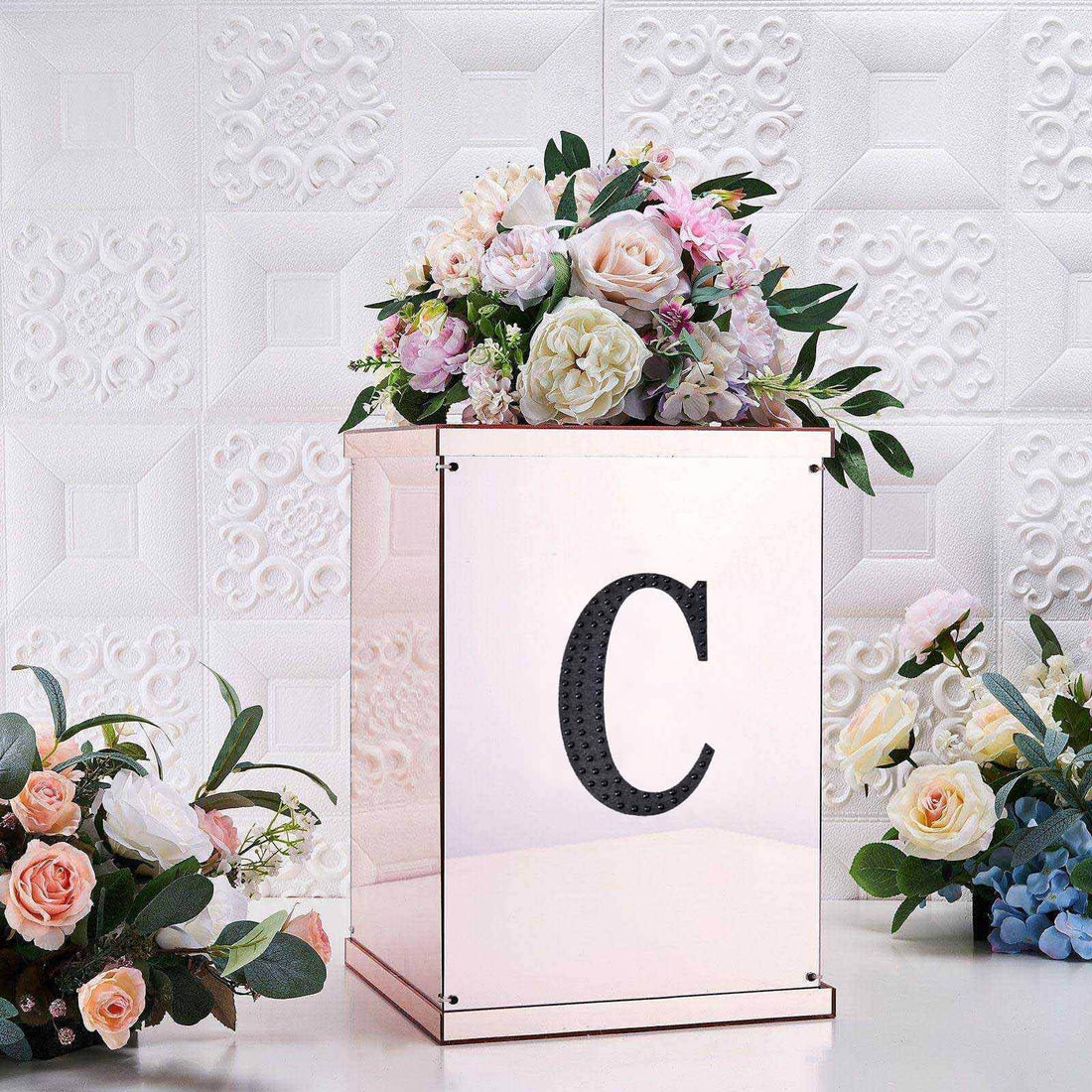 Elegant floral arrangement in a mirrored box with letter "C" on a textured backdrop.