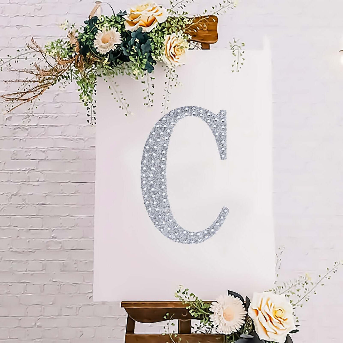 Glittery letter "C" on a decorated canvas with flowers and greenery.