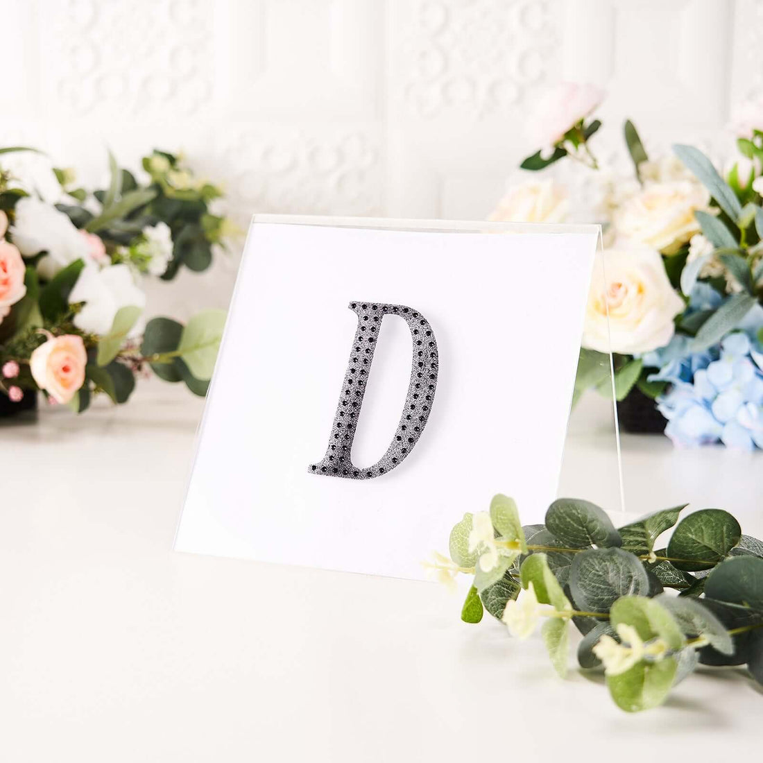 Decorative letter "D" in rhinestones on display with floral backdrop.