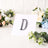 Decorative letter "D" in rhinestones on display with floral backdrop.