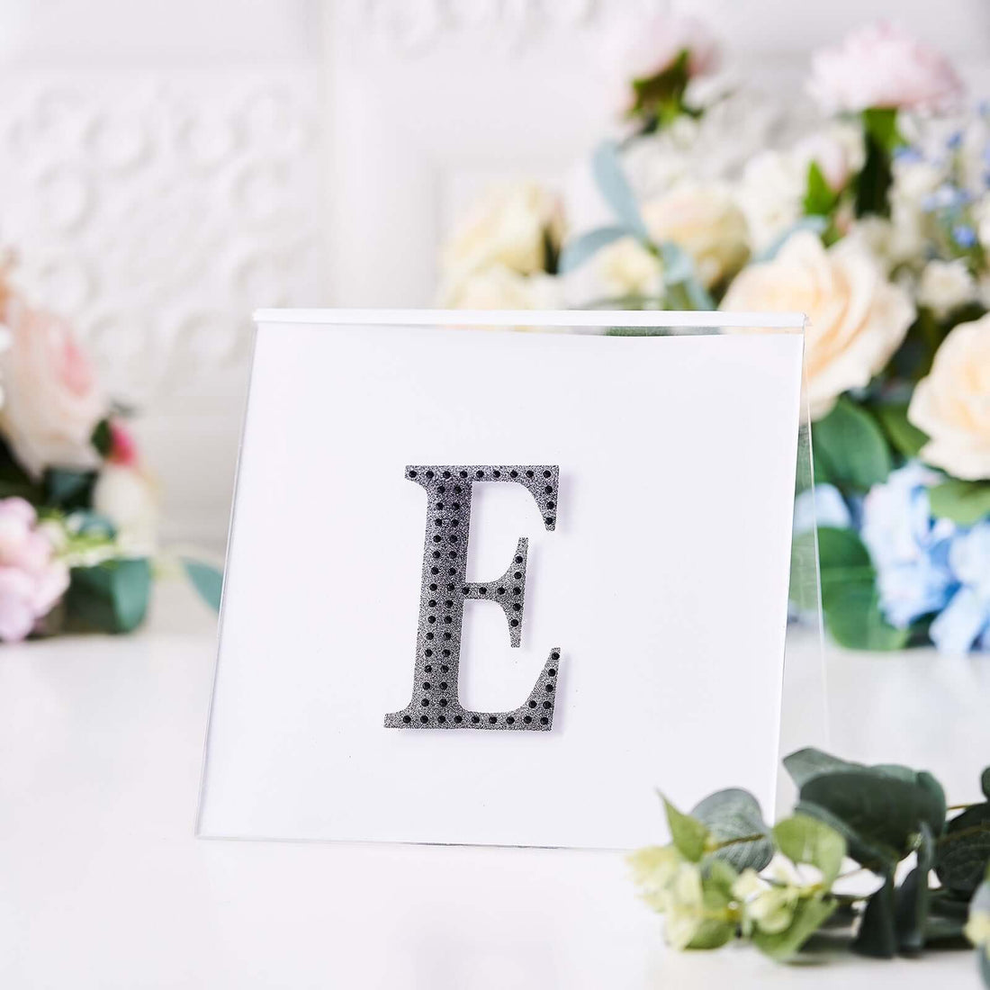 Decorative letter 'E' displayed on a clear acrylic stand with floral background.