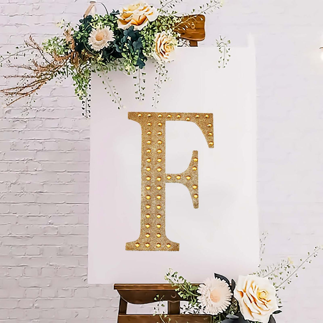 Gold letter "F" decorative sign with lights on an easel surrounded by flowers.