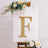 Gold letter "F" decorative sign with lights on an easel surrounded by flowers.