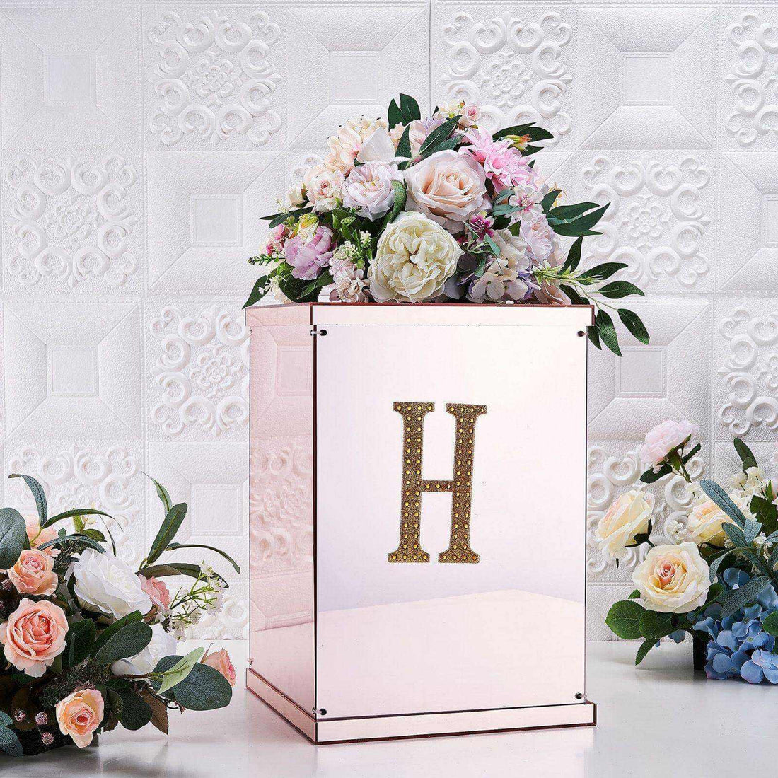 Rose gold decorative box with floral arrangement and letter "H" on a textured background.