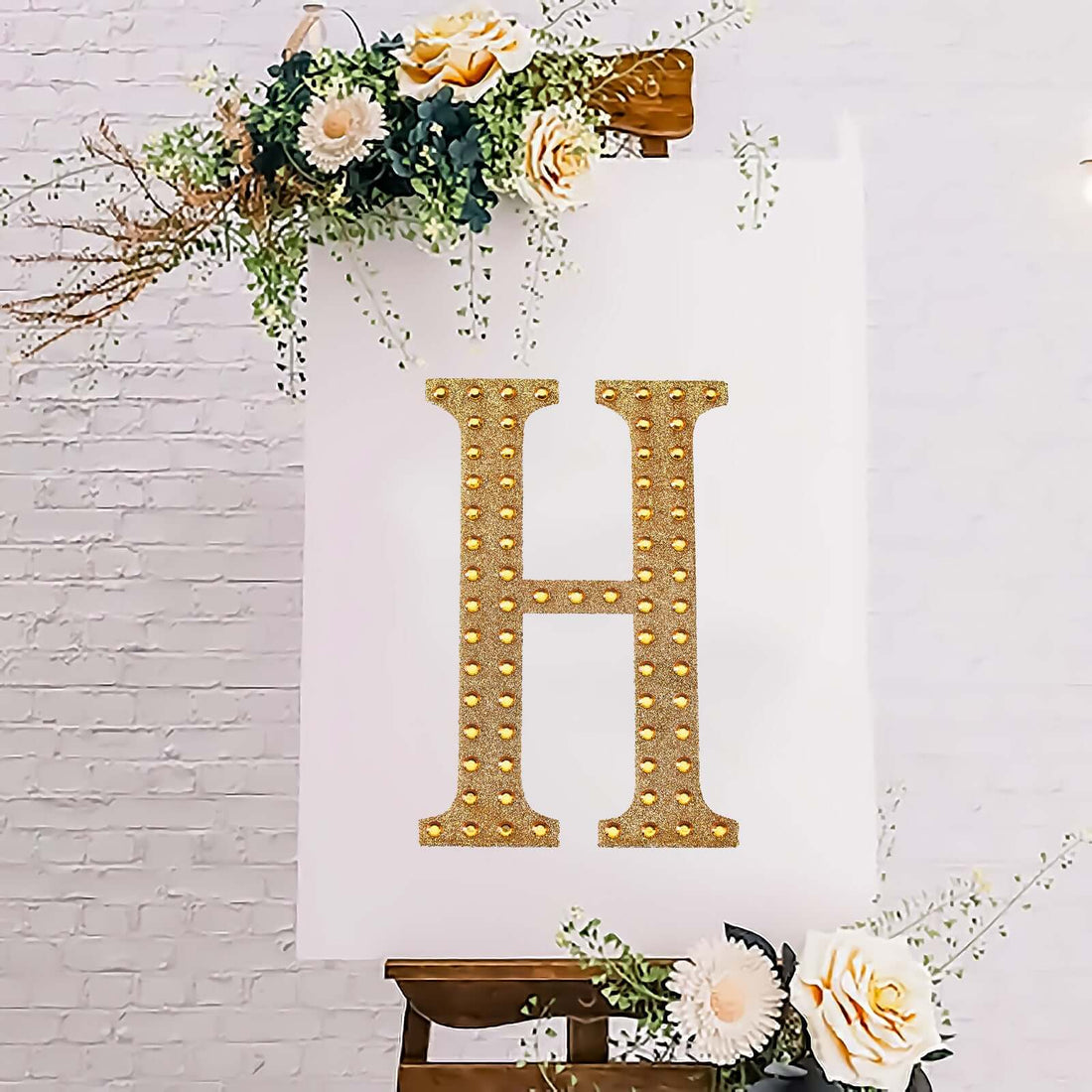 Gold marquee letter "H" adorned with flowers on an easel backdrop.