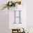 Silver glitter letter "H" on a decorative easel with flowers.