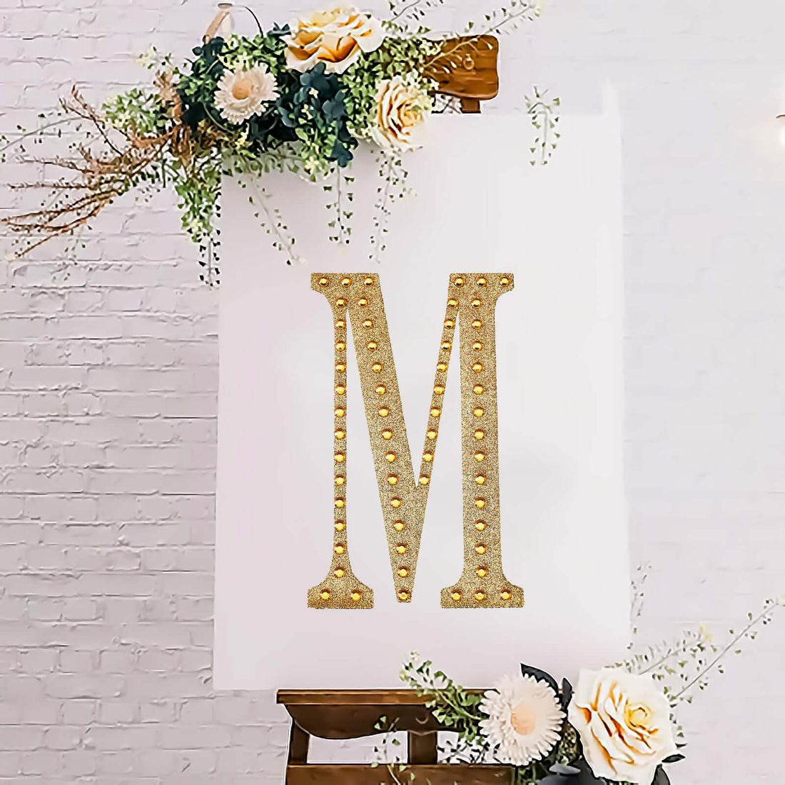Gold glitter letter "M" adorned with flowers on easel backdrop.