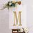 Gold glitter letter "M" adorned with flowers on easel backdrop.