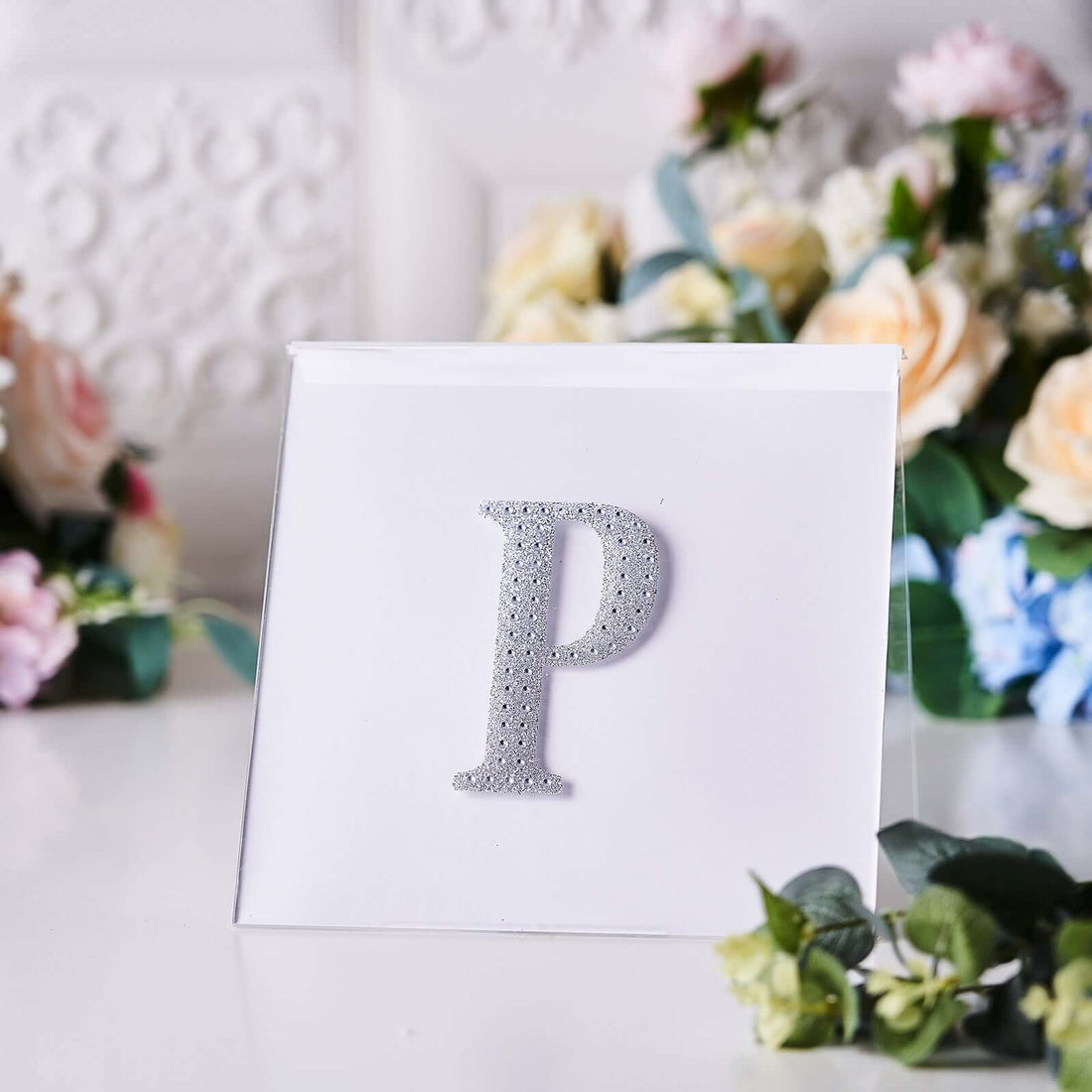 Glittery silver letter "P" on a white card surrounded by flowers.