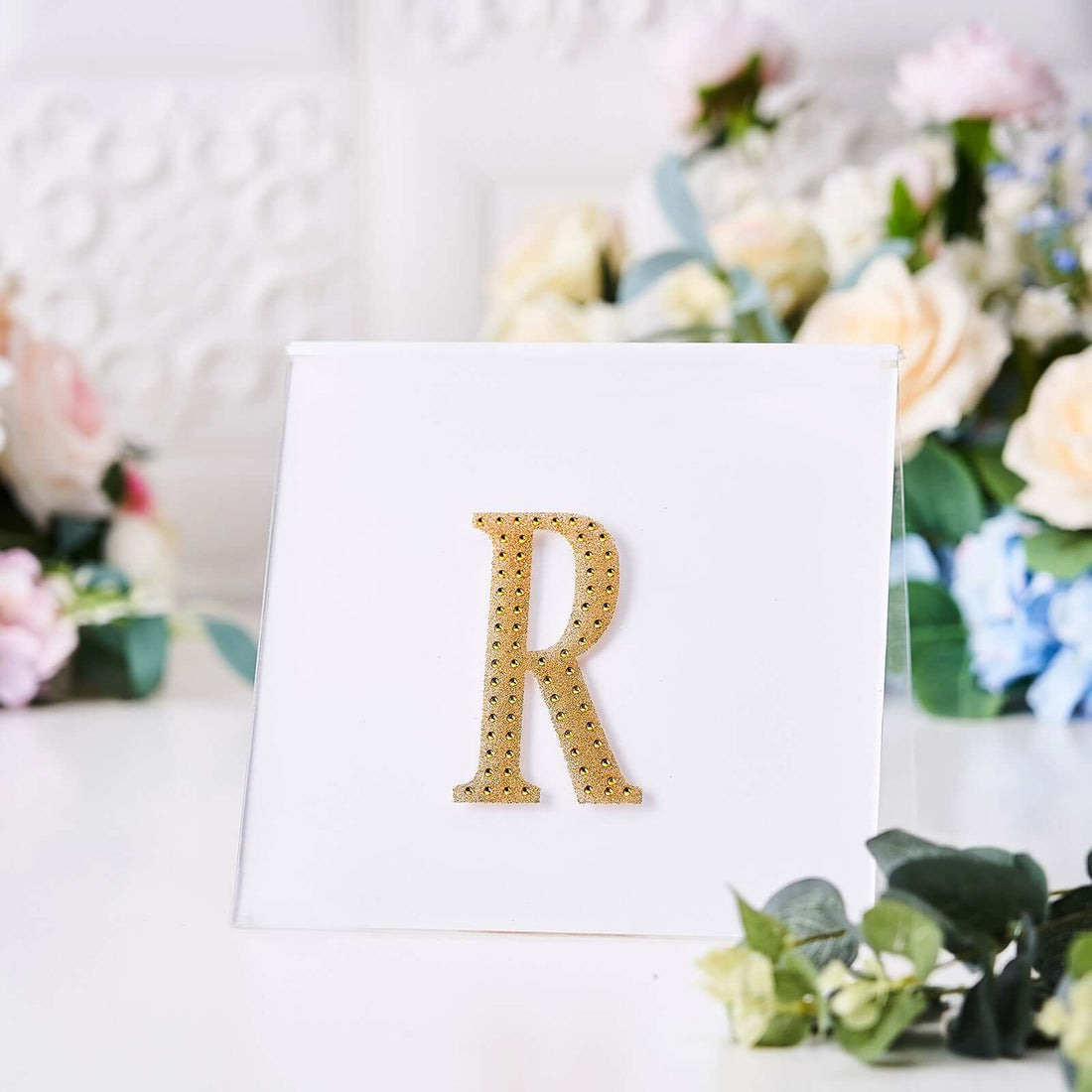 Gold glitter letter "R" on a clear display stand with floral background.