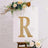Gold rhinestone letter R on an easel surrounded by flowers.