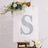 Decorative letter "S" with floral accents on an easel in a styled setting.
