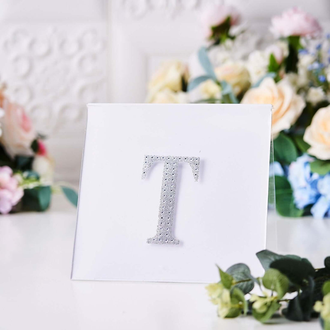 Sparkling letter T decoration on a white card with floral background.