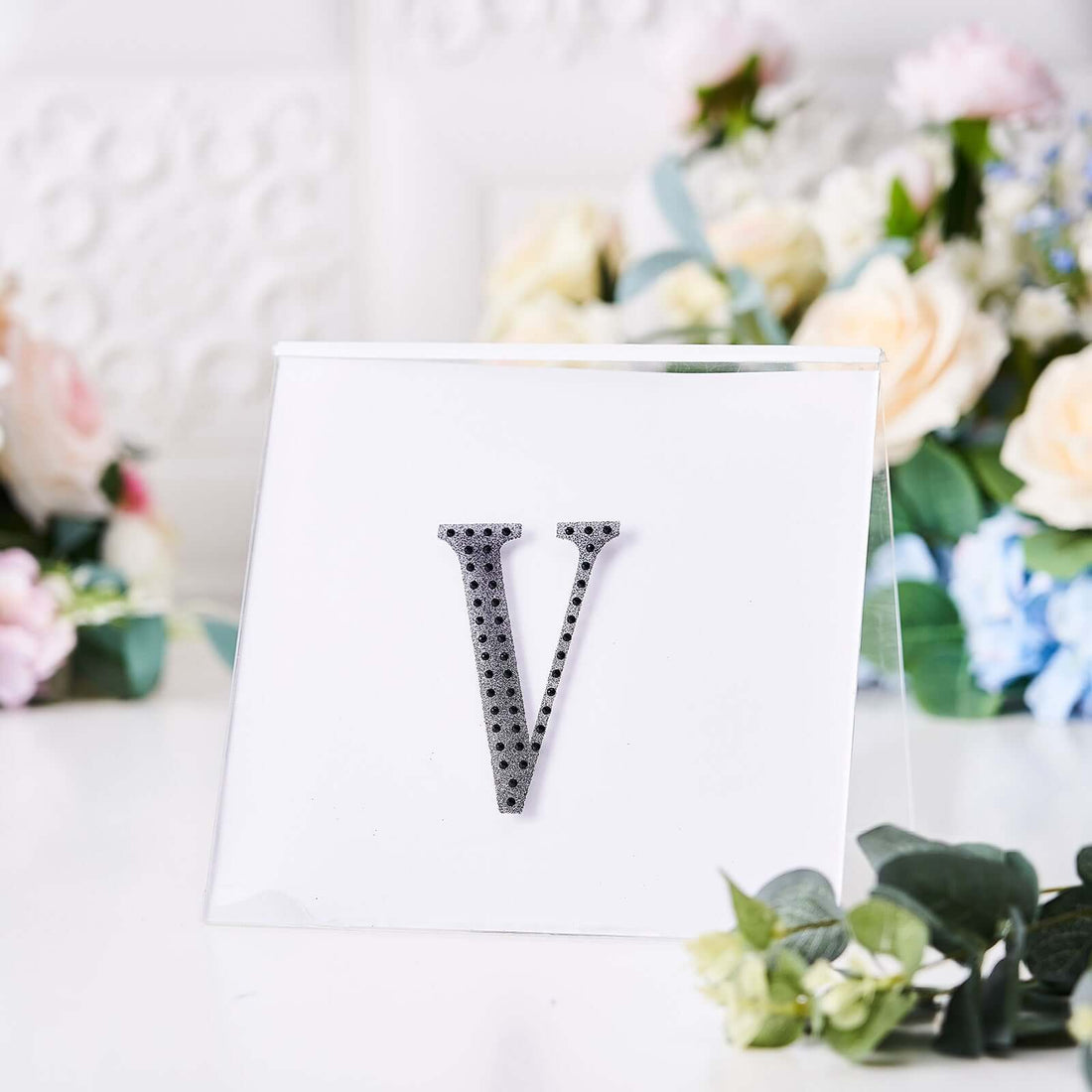 Stylish decorative letter "V" on a clear stand with floral backdrop.