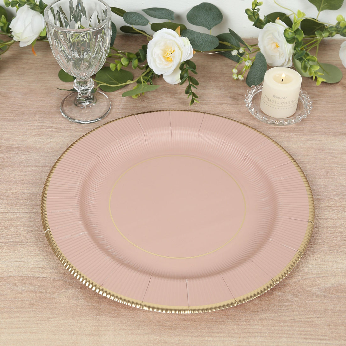 Pink decorative plate with gold trim on a wooden table with greenery and candles.