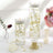 Elegant decorative candles in glass vases with gold accents and floral backdrop.