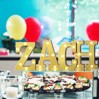 Gold marquee letters spelling "ZACH" at a festive dessert table with colorful balloons.