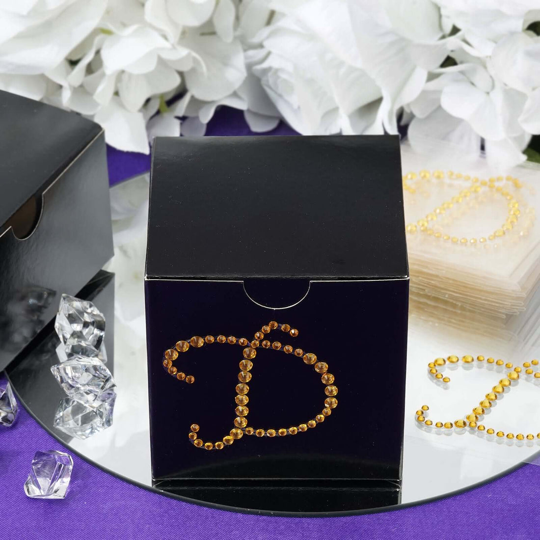 Black gift box with rhinestone letter "D" design, surrounded by decorative elements.