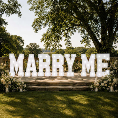 Illuminated MARRY ME proposal sign with white roses in a garden setting