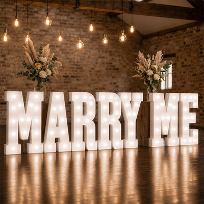 Illuminated MARRY ME wedding proposal sign with floral arrangements in rustic venue