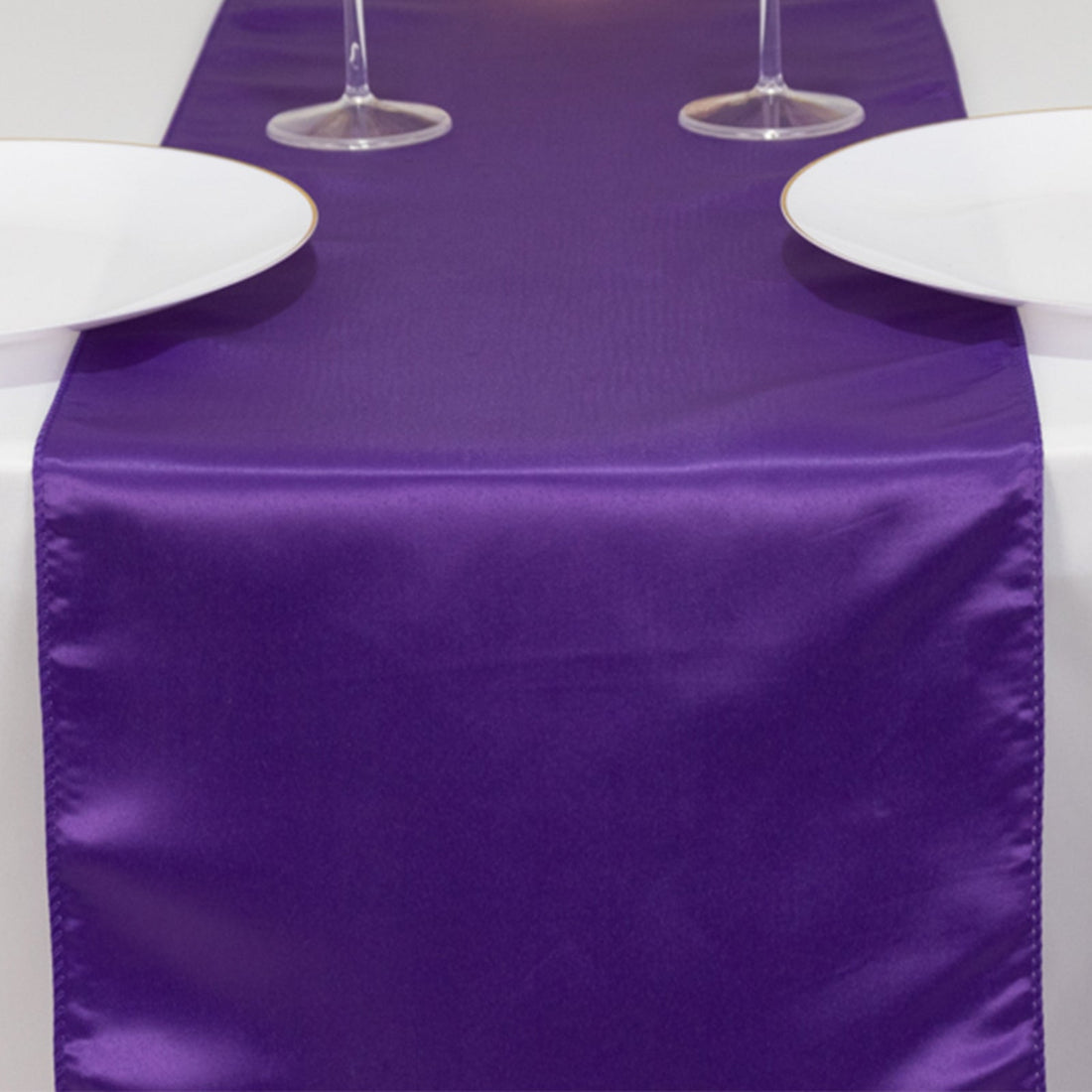 Purple satin table runner on a white table with plates and glasses.