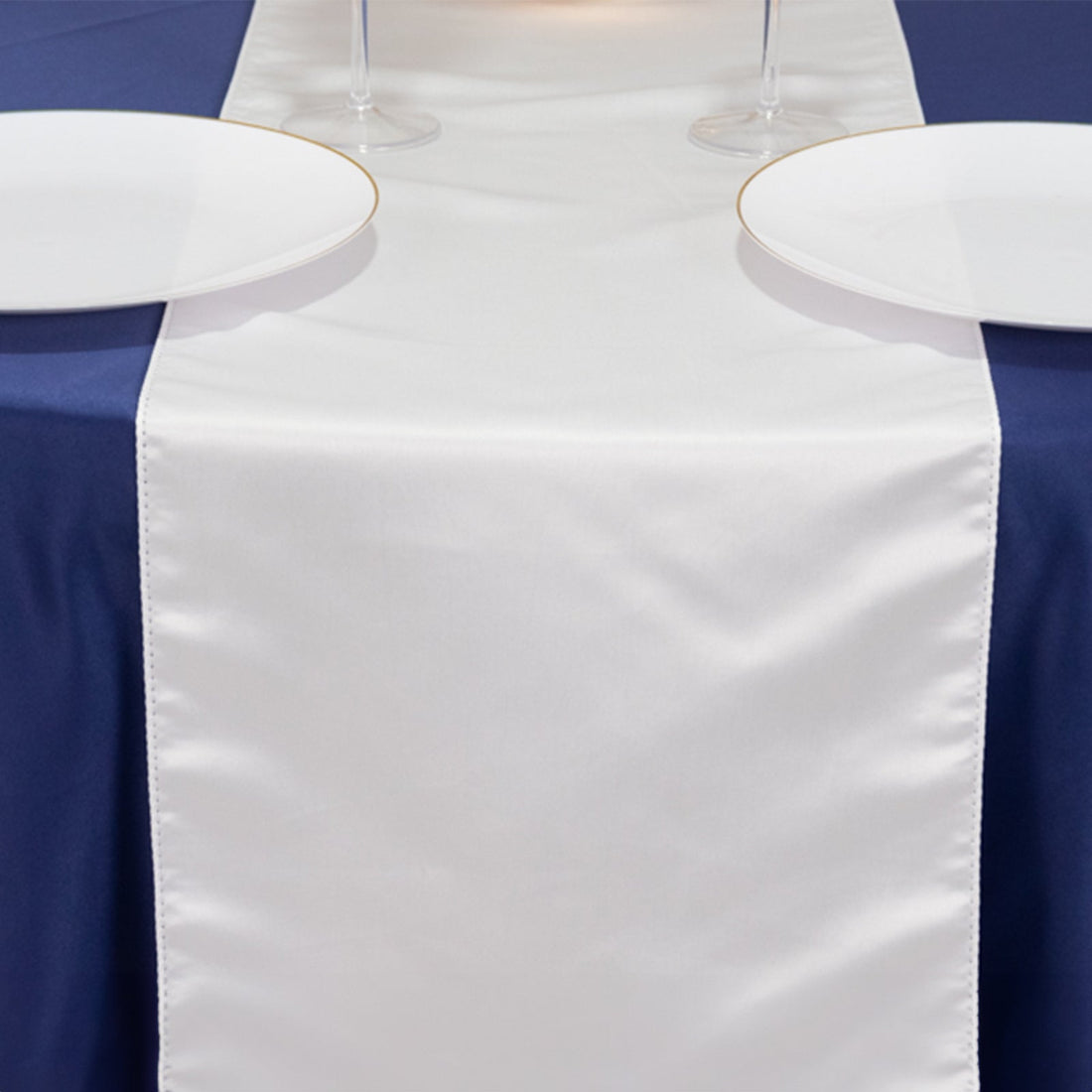 White satin table runner on a navy tablecloth with plates and glasses.