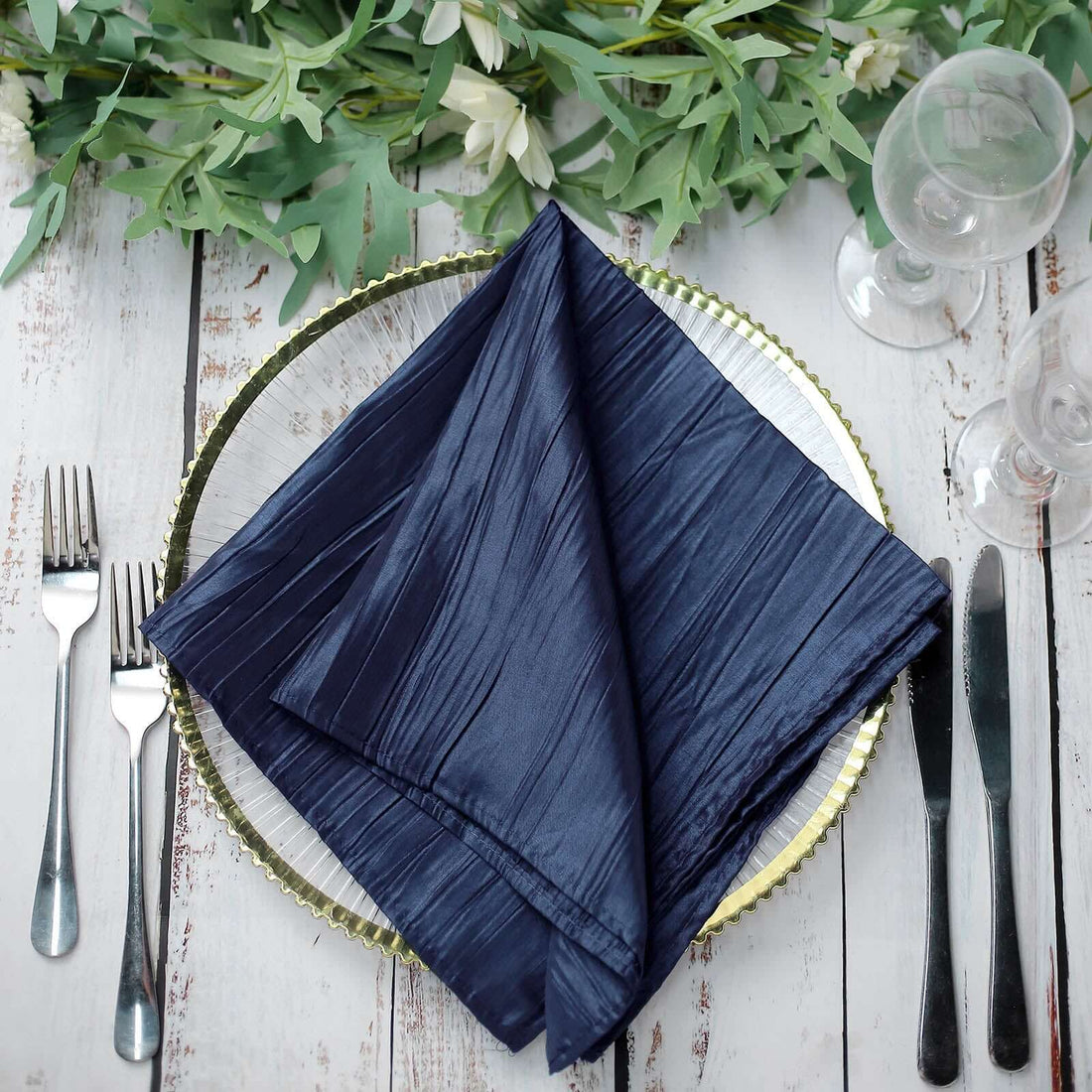 Navy blue pleated napkin on a silver charger plate with cutlery and greenery #whtbkgd