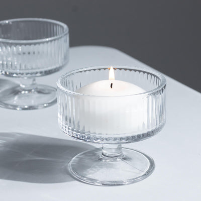 Lit white candle in a clear glass pedestal holder on a white surface