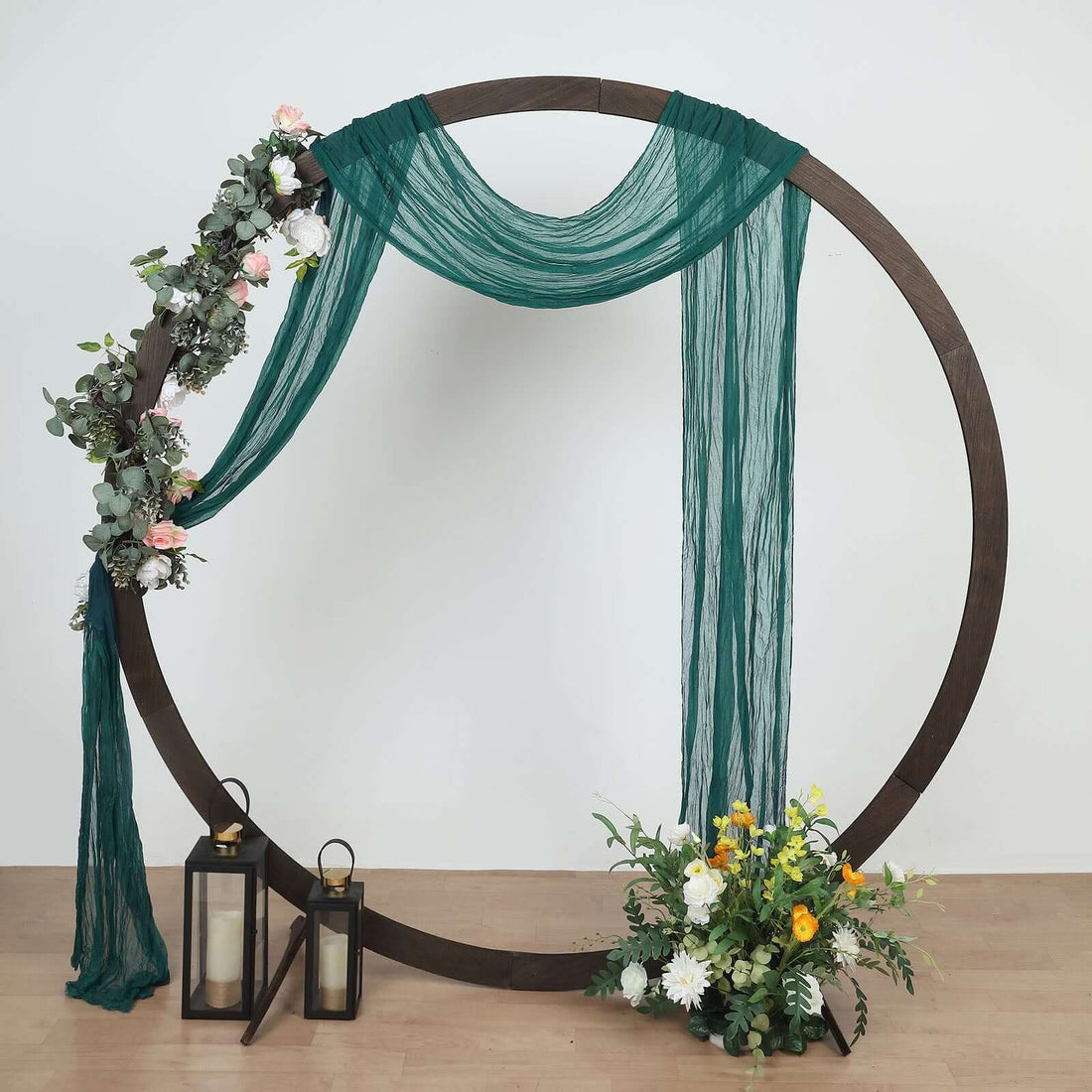 Decorative floral arch with draped fabric and lanterns, set on a wooden floor.