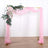Pink draped wedding arch with floral accents on a plain backdrop.