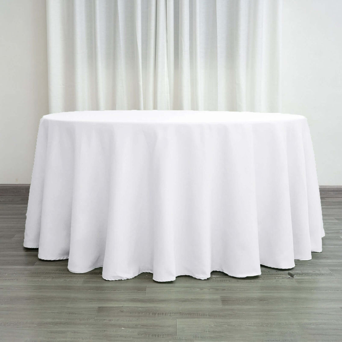 Round white tablecloth on a wooden floor with curtains in the background.
