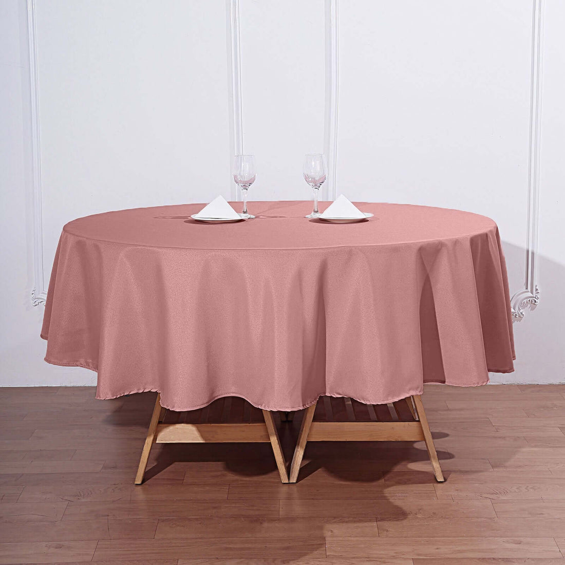 Round pink tablecloth draped over a wooden table set with glasses and napkins.