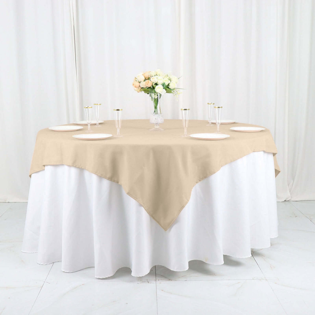 Beige tablecloth draped over a round table with white undercloth and floral centerpiece.