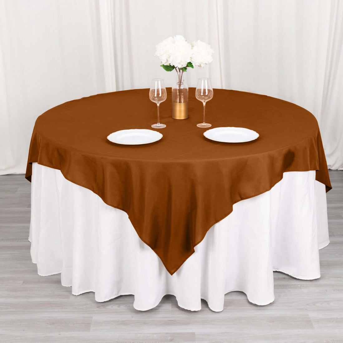 Brown round tablecloth draped over a white table with plates and glasses.