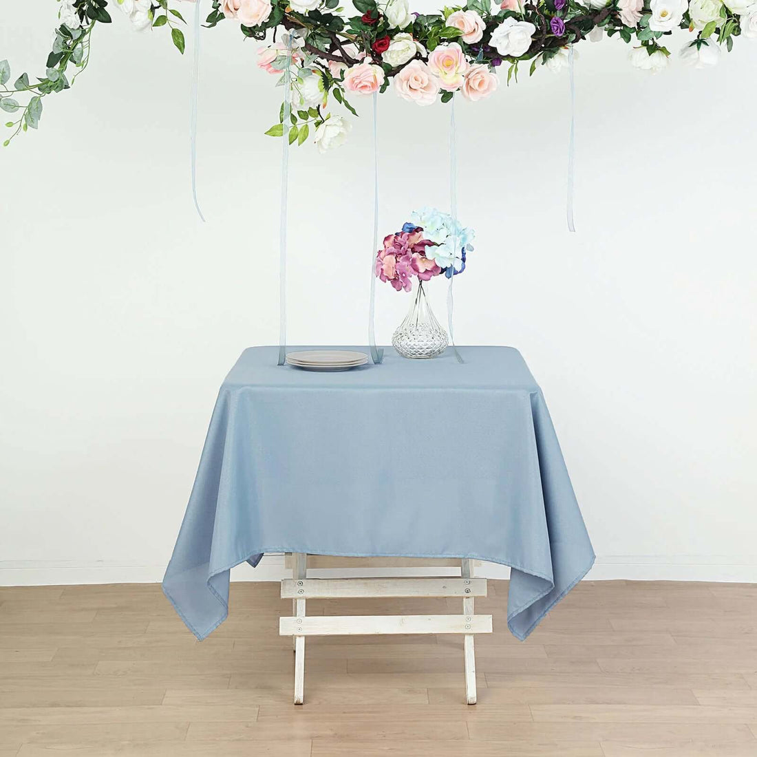 Blue tablecloth on a wooden table with flowers above.