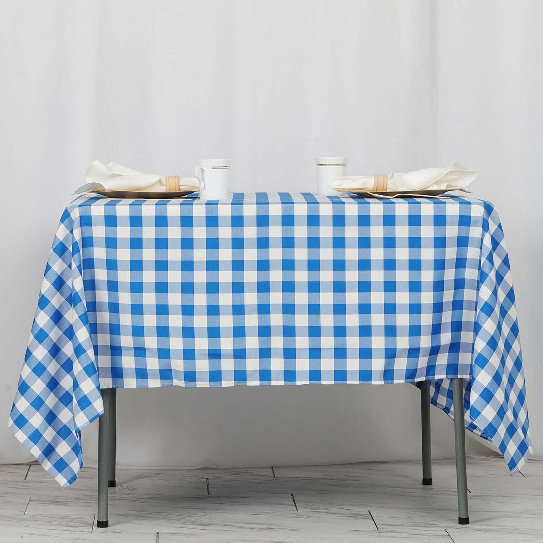 Blue and white checkered tablecloth on a table with cups and napkins.