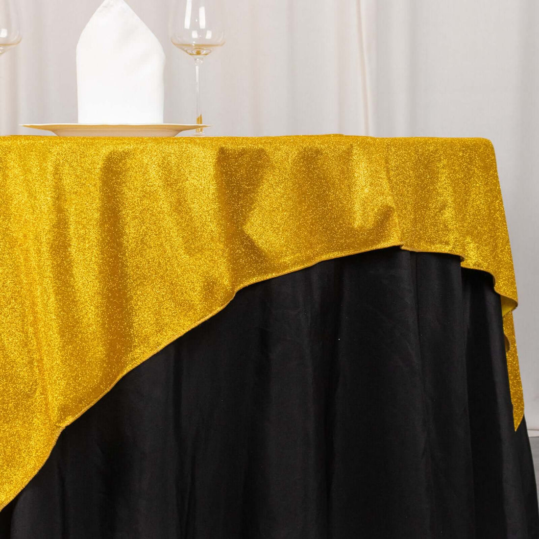 Gold glitter tablecloth draped over a black table with napkin and glasses.
