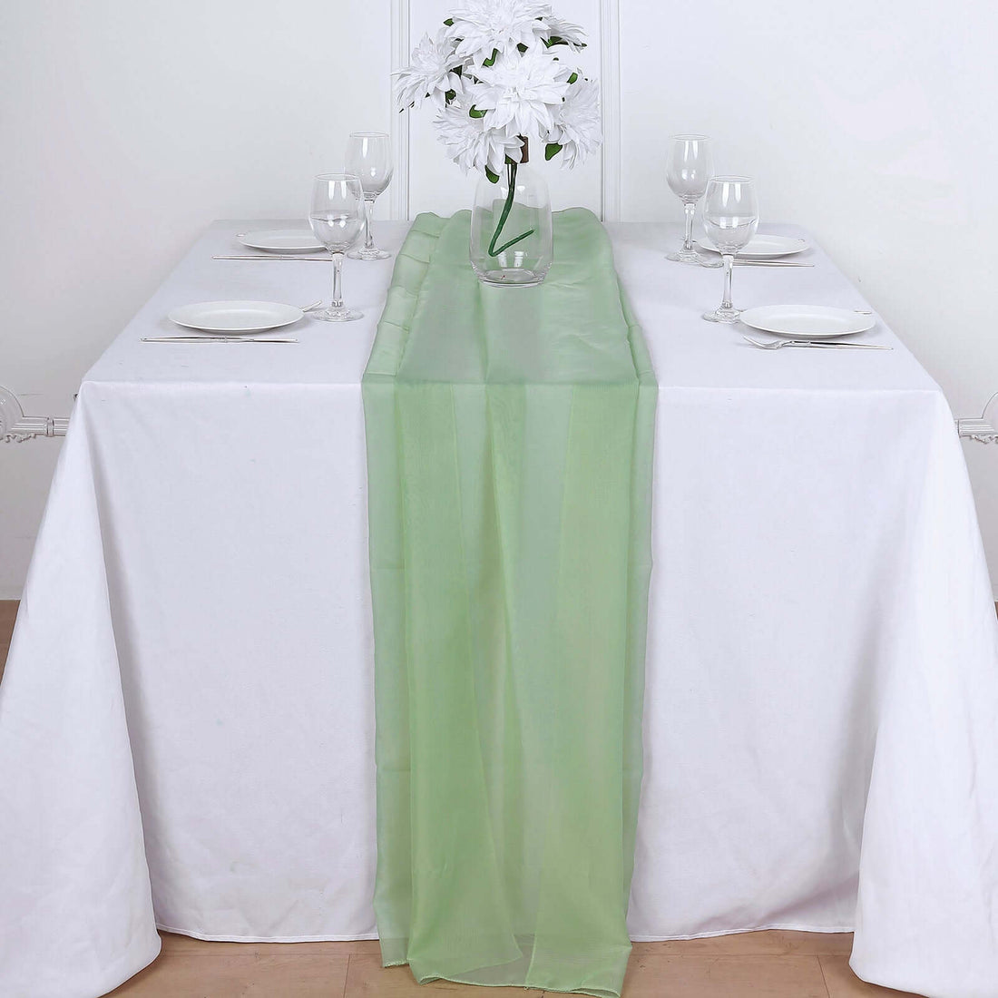 Green table runner on a white table set for dining with flowers and glassware.